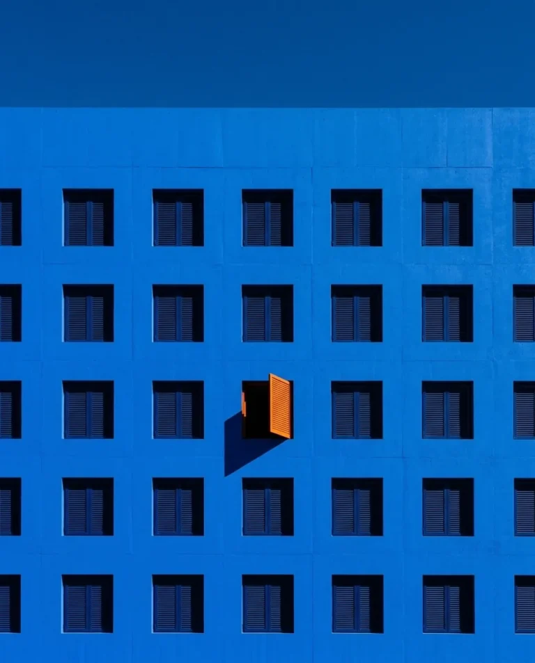 Fachada azul con una retícula de ventanas y una sola contraventana naranja abierta
