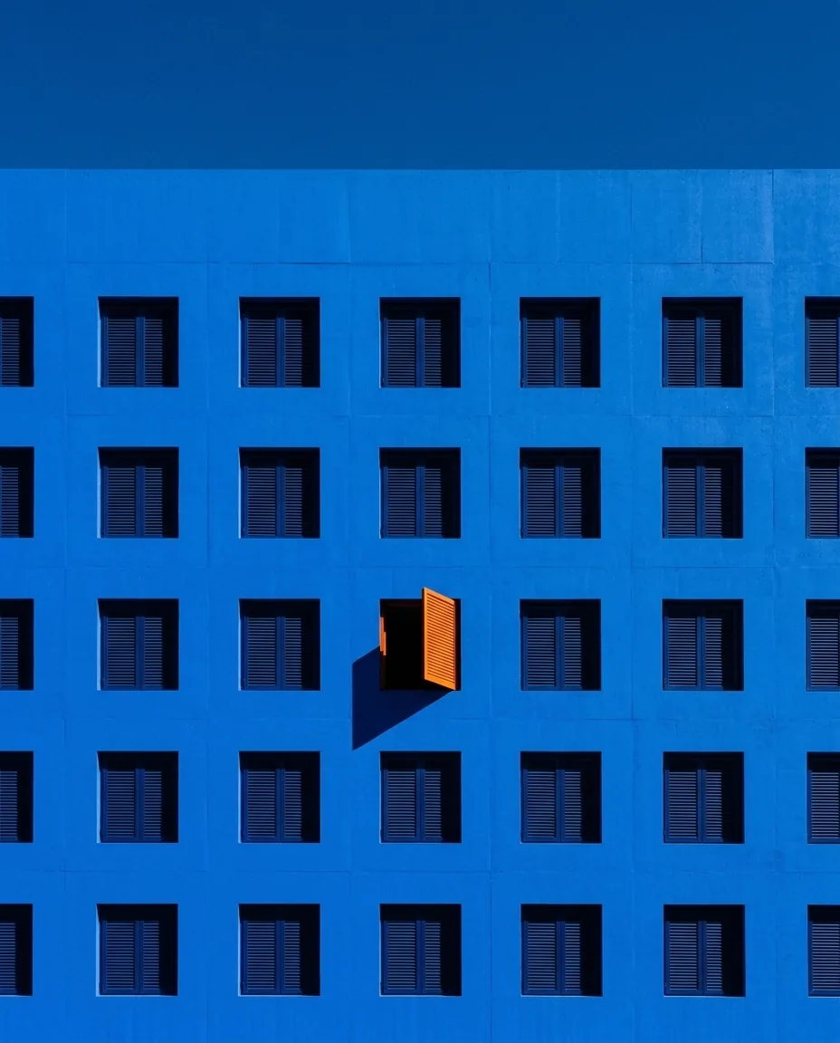 Fachada azul con una retícula de ventanas y una sola contraventana naranja abierta
